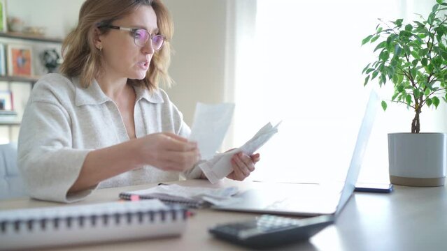 Beautiful middle-aged woman in glasses extremely surprised with higher expenses and throwing bills at laptop at home office. Small business troubles, home utilities, money savings, Mortgages 4K video.