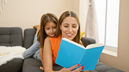 Fototapeta na wymiar A smiling woman and her daughter enjoying reading a blue book together on a cozy sofa in their living room.