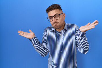 Hispanic man with beard wearing glasses clueless and confused expression with arms and hands...