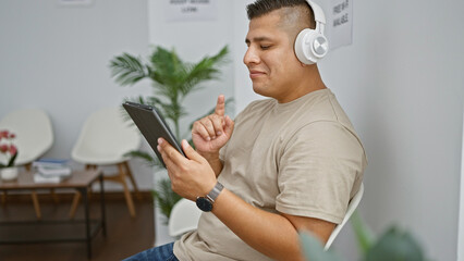 Chill young latin man enjoying catchy songs with headphones, sitting comfortably in a waiting room, confidently smiling at camera on his touchpad