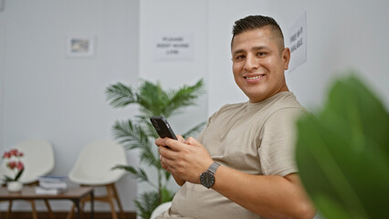 Cheerful young latin man happily texting away on his smartphone while sitting comfortably in a waiting room chair