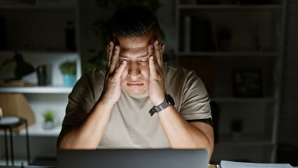 Stressed latin man tired of overwork at his desk, seriously tackling business hurdles with laptop...