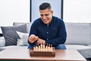 Young latin man playing chess sitting on sofa at home