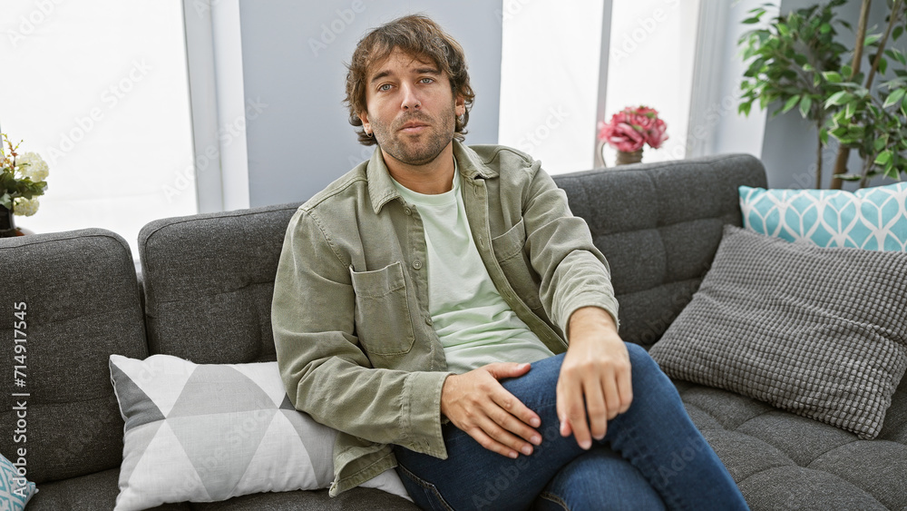 Poster a relaxed young man with a beard sits comfortably on a sofa in a modern living room setting.