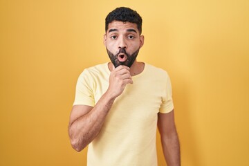 Hispanic man with beard standing over yellow background looking fascinated with disbelief, surprise and amazed expression with hands on chin