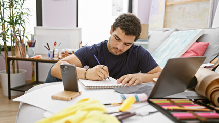 A young bearded man studying with a laptop in an artistically cluttered indoor environment.