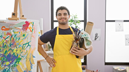 A smiling young man in a yellow apron holding paintbrushes stands before an abstract painting in an...