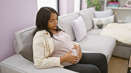 Fototapeta premium Serious-faced, pregnant young woman sitting at home, casually touching her belly on her living room sofa, embodying the emotion and relaxed anticipation of motherhood.