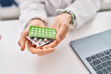Young caucasian woman doctor holding birth control pills sitting on table at clinic