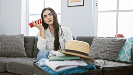 Attractive hispanic woman packing suitcase in living room, contemplating with smartphone in hand.