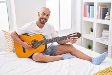 Young bald man playing classical guitar sitting on bed at bedroom