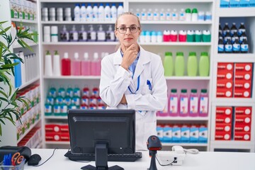 Young caucasian woman working at pharmacy drugstore looking confident at the camera smiling with crossed arms and hand raised on chin. thinking positive.