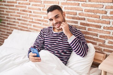 Young hispanic man using smartphone sitting on bed at bedroom