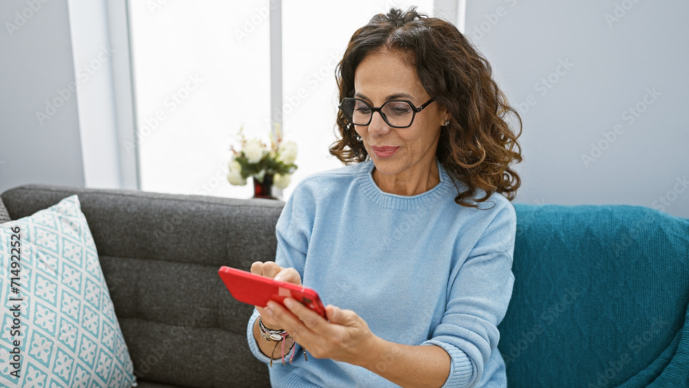 Canvas Prints a mature woman with glasses and curly hair focuses on her smartphone while sitting on a gray sofa in