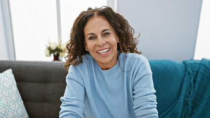 Smiling middle-aged hispanic woman with curly hair sitting on a couch in a cozy living room.