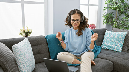 Hispanic woman enjoying music with headphones in a cozy living room with plants and natural light.