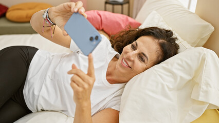 Obraz premium Smiling middle-aged hispanic woman using smartphone in a cozy bedroom setting.