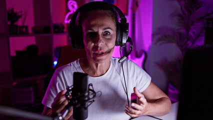Mature woman with headset streaming in a neon-lit gaming room at night, expressing excitement.