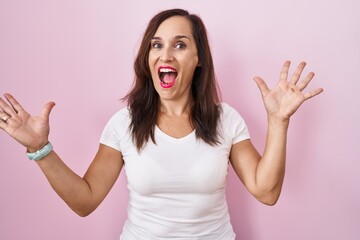 Middle age brunette woman standing over pink background celebrating crazy and amazed for success with arms raised and open eyes screaming excited. winner concept