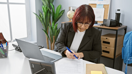 Hispanic senior woman focused on signing a document in a modern office workplace setting.