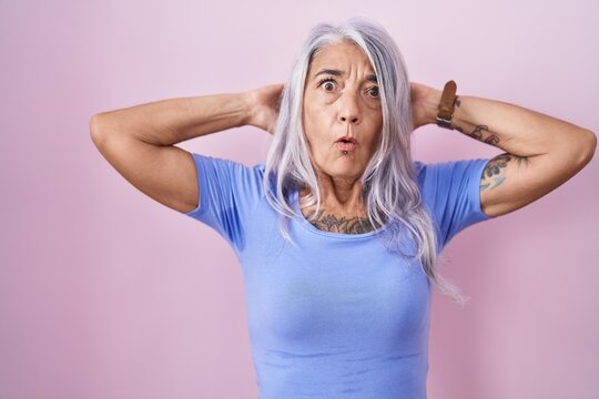 Middle age woman with tattoos standing over pink background crazy and scared with hands on head, afraid and surprised of shock with open mouth