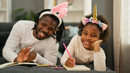 African american father and daughter wearing funny diadem lying on sofa drawing at home