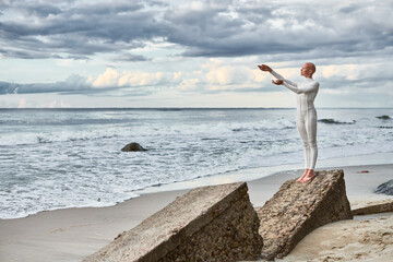 Full length portrait of hairless girl with alopecia in white futuristic suit standing on stone sea...