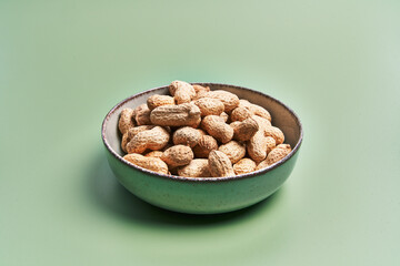 A bowl filled with unshelled peanuts presented on a plain green background, depicting simplicity and organic food concepts.