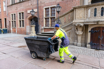 
A worker pushes a wastebasket on a city street against a background of buildings