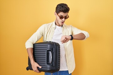 Young hispanic man holding suitcase going on summer vacation looking at the watch time worried,...