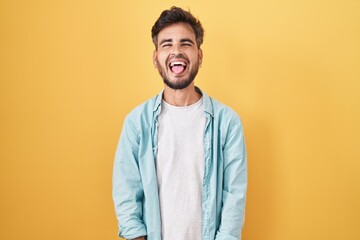 Young hispanic man with tattoos standing over yellow background sticking tongue out happy with...
