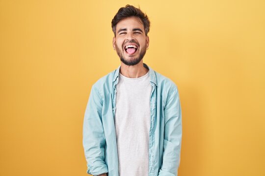 Young hispanic man with tattoos standing over yellow background sticking tongue out happy with funny expression. emotion concept.