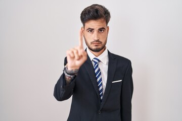 Young hispanic man with tattoos wearing business suit and tie pointing with finger up and angry expression, showing no gesture