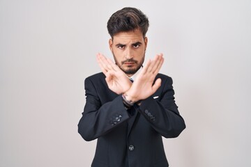 Young hispanic man with tattoos wearing business suit and tie rejection expression crossing arms and palms doing negative sign, angry face