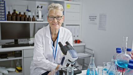 Fototapeta premium Confident, smiling senior woman scientist with grey hair, happy at her work, masterfully using microscope in lab, submitting science to her mature wisdom in medical research center.
