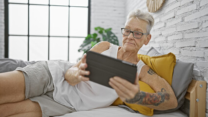 Fototapeta premium Serious mature grey-haired woman relaxing, fully engrossed in her technicolor world online, lying comfortably amidst the pillows in her homely bedroom, screen lit face peeking from cosy blanket.