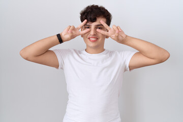 Young non binary man wearing casual white t shirt doing peace symbol with fingers over face, smiling cheerful showing victory