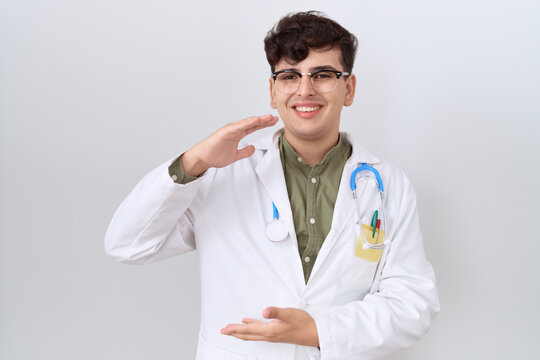 Young non binary man wearing doctor uniform and stethoscope gesturing with hands showing big and large size sign, measure symbol. smiling looking at the camera. measuring concept.