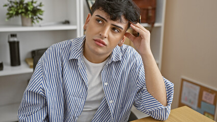 Pensive young man in office setting with striped shirt touching hair and contemplative look.