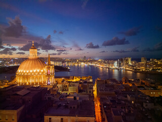 Fototapeta premium Beautiful illuminated dome of Basilica of Our Lady of Mount Carmel in Valletta. Aerial view with bright orange street line towards Sliema skyline and light reflection in the sea.