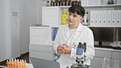 A young man wearing a lab coat sits contemplatively in a modern laboratory filled with equipment and chemistry bottles.