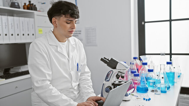 A young man in a white lab coat works intently on a laptop in a bright laboratory, with a microscope and scientific equipment around.