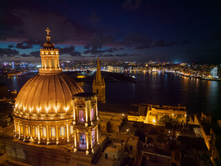 Obraz premium Basilica of Our Lady of Mount Carmel at dusk in historic Valletta city. Aerial view of illuminated dome and the coastline of Sliema in Malta.