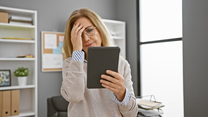 Worried middle-aged woman in office looking at tablet with hand on forehead evoking concern or confusion.