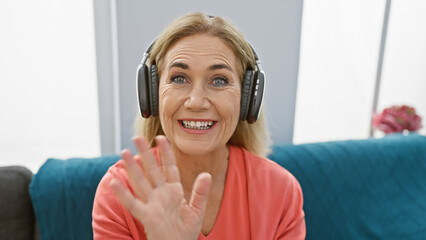 A smiling caucasian woman wearing headphones gestures in a modern living room.