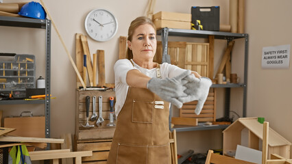 Mature woman in apron stretching gloves in a woodworking studio indicates safety and skill.
