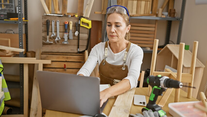 A mature woman in a woodworking studio focuses intently on her laptop amidst various carpentry tools.