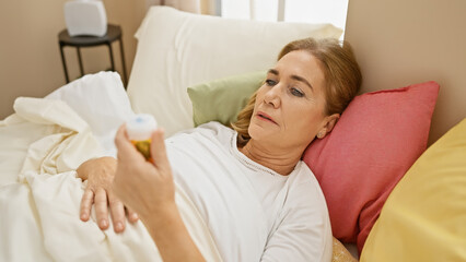 A senior caucasian woman is lying in bed looking at a pill bottle in her home bedroom, suggesting...