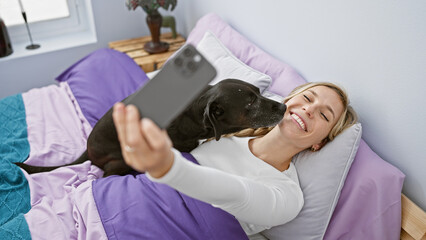 Blonde woman takes a selfie with her black labrador in a cozy bedroom setting.