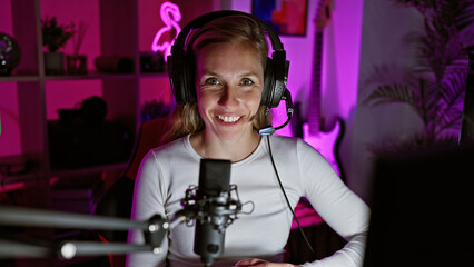 Smiling woman with headphones in a neon-lit gaming room with a microphone and computer at night.
